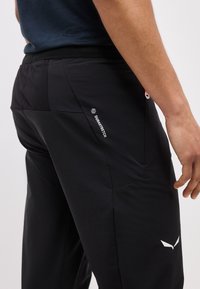 Άνδρας που φοράει μαύρο παντελόνι από ύφασμα Durastretch με μια μικρή τσέπη από δίχτυ και λευκό λογότυπο στο μηρό, στέκεται μπροστά σε απλό φόντο.