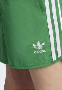 Zelené šortky vyrobené z lehkého materiálu s bílým trojitým pruhem a vyšitým logem Adidas na levé noze. Hladký povrch.