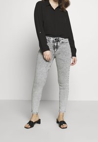 Camisa negra de manga larga con botones, jeans ajustados grises claros con una textura desvanecida y sandalias negras de tiras con tacón cuadrado.