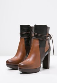 Bruine en zwarte leren ankle boots met hoge hak en een zijrits, versierd met een wikkeldraaddetail, op een witte ondergrond.