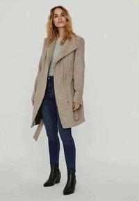 Kvinne som har på seg en beige lang frakk over grå skjorte, mørkeblå skinny jeans og svarte ankelstøvler, stående mot en ensfarget lys bakgrunn.