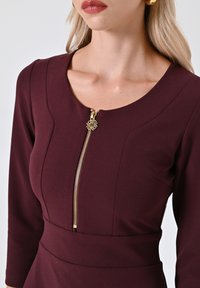 Abito bordeaux con scollatura a barca e maniche a tre quarti, caratterizzato da un dettaglio con zip dorata e vita sagomata. Tessuto con una trama liscia.