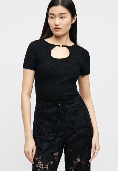 Νεαρή γυναίκα με μακριά μαύρα μαλλιά που φοράει μαύρο ριμπ κοντομάνικο τοπ με άνοιγμα-κλειδί στο στήθος και μαύρο παντελόνι από διακοσμητική δαντέλα.