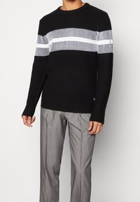 Musta ribbineuloksinen pusero, jossa on valkoiset vaakaraitakuviot ja teksturoitu kangas. Pyöreä pääntie, pitkät hihat ja rento istuvuus.