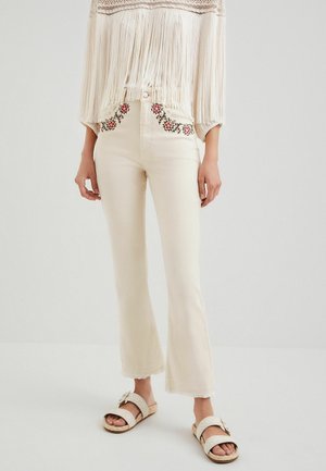 Femme portant un jean taille haute écru avec broderies florales près des poches, un haut crème à franges et des sandales beige à boucles, debout sur un sol blanc.