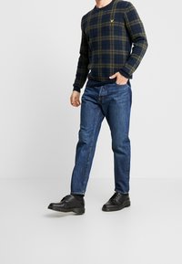 Pull en tartan navy avec des accents verts, poignets et ourlet côtelés ; porté avec un jean en denim bleu et des chaussures à lacets noires sur un fond clair.