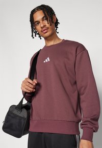Bordeaux sweatshirt gemaakt van zachte stof met een ronde halslijn en geribde manchetten. Heeft een klein wit grafisch logo op de borst. Zwarte tas met mesh accenten.