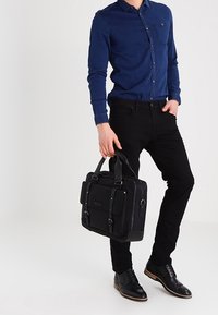 Svart portfölj med dubbla bärremmar, framflik och spännelement; kombinerad med en blå skjorta med knappar och svarta byxor. Utstrålar en snygg design.