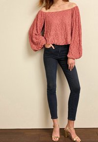 Rosa, tekstureret off-the-shoulder top med lange, voluminøse ærmer; parret med mørke skinny jeans og gyldne hæle sandaler.