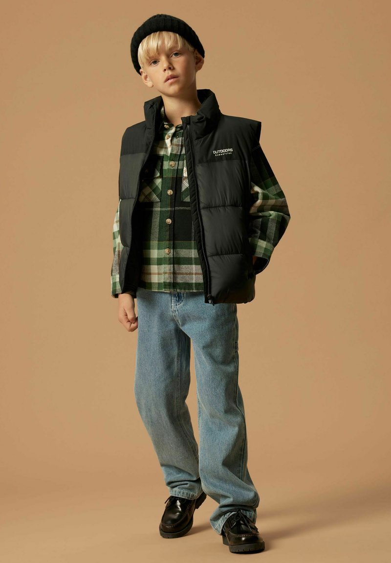 Jongen in een zwarte beanie, zwarte gewatteerde vest, groen geruiten shirt, lichtblauwe spijkerbroek en zwarte schoenen, staand tegen een beige achtergrond.