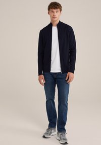 Marinblå zip-jacka över en vit t-shirt, kombinerat med blå jeans och grå sportskor. Modellen står framför en neutral bakgrund.
