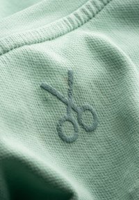 Tessuto verde menta con una consistenza morbida, caratterizzato da un'icona di forbici ricamata in una tonalità di verde leggermente più scura. Le cuciture sono ben rifinite.