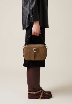 Brown textured leather handbag with a top handle and a gold circular detail on the front, held by a person wearing a black outfit and brown shoes.