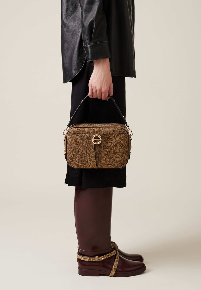 Brown textured leather handbag with a top handle and a gold circular detail on the front, held by a person wearing a black outfit and brown shoes.