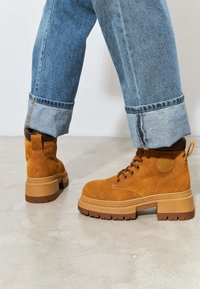 Bruine suède enkellaarsjes met een stevige rubberen zool, vetersluiting aan de voorkant en een cirkelvormig logo aan de zijkant. Er zijn spijkerbroeken met opgerolde manchetten zichtbaar.
