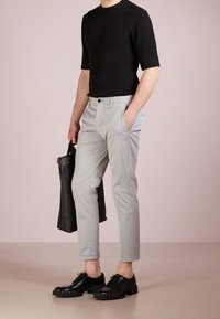 Black ribbed short-sleeve top, light grey checkered trousers, black lace-up shoes with stud details, holding a black leather bag.