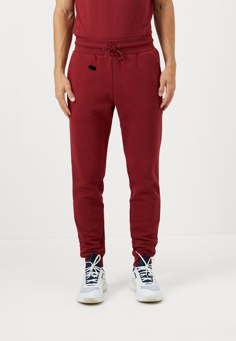 Burgonya színű melegítőnadrág, húzózsinórral a deréknál, keskeny lábszárral és egy kis fekete díszítő zsebel. Puha, texturált anyagból készült.