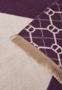 Gros plan sur un tissu tissé avec des franges beiges, présentant un motif géométrique violet superposé sur un fond beige et violet.