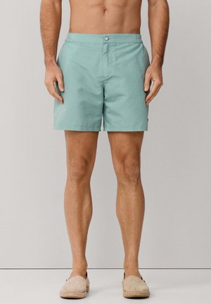 Muž oblečený v svetlomodrých šortkách a béžových šmykľavých topánkach stojaci pred jednoliatym svetlosivým pozadím.