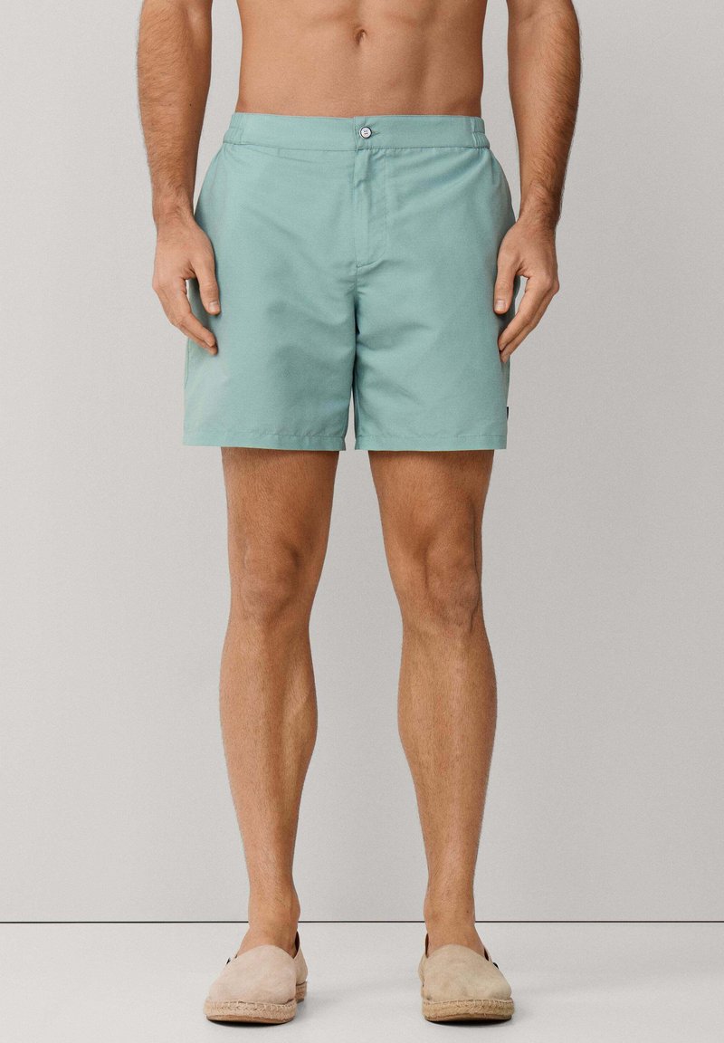 Hombre con pantalones cortos azul claro y zapatos beige sin cordones, de pie frente a un fondo liso gris claro.