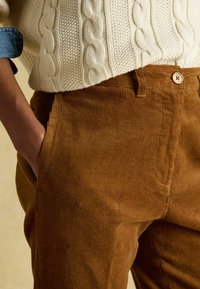 Brūnas kordurojas bikses ar smalkām vertikālām svītrām. Tām ir pogas aizdare un sānu kabatas. Kopā ar tamborētu krēmkrāsas džemperi.