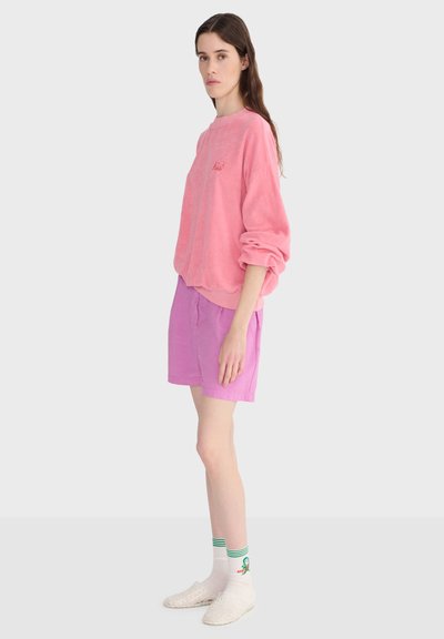 Jeune femme de profil portant un sweat-shirt rose ample, un short lilas, des chaussettes blanches à rayures vertes et des chaussures blanches sans lacets sur un fond uni.