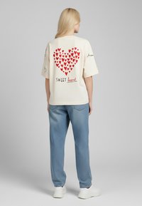 T-shirt crème à manches courtes avec un motif de cœurs rouges au dos, comportant le texte "SWEET" en dessous, associé à un jean bleu clair et des baskets blanches.