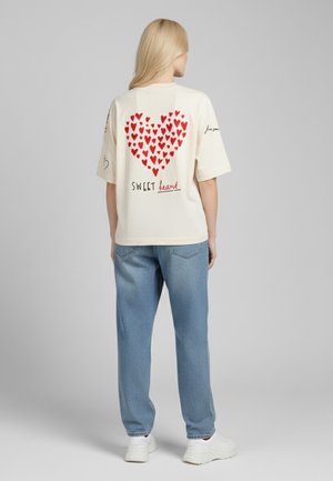 Crème T-shirt met korte mouwen en een rood hartpatroon op de achterkant, met daaronder de tekst "SWEET", gecombineerd met lichtblauwe jeans en witte sneakers.