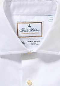 Wit overhemd met een puntkraag en een enkele gele knop. Etiket leest "Brooks Brothers Luxury Shirt Collection," gemaakt van Italiaanse stoffen.