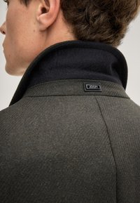 Donkergroene gelaagde jas met een hoge kraag, voorzien van een marineblauwe voering en een zwart merklabel nabij de kraagnaad.