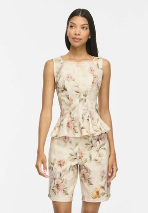 Donna che indossa un top floreale color crema senza maniche con vita peplo e pantaloncini abbinati al ginocchio su uno sfondo bianco semplice.