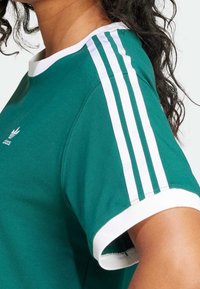 Persona que lleva una camiseta verde de Adidas con tres rayas blancas en el hombro y ribete blanco en la manga.
