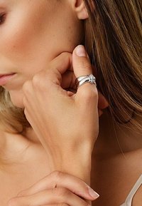 Sølvfarvet ring med sammenflettede bånd, der fremhæver en poleret overflade og en glat tekstur. Båret på fingeren af en hånd mod hud.