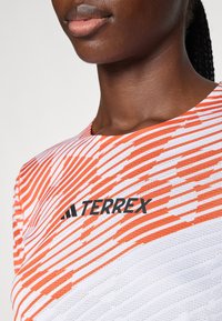 Červený a bílý pruhovaný atletický top s texturovanou tkaninou; má výrazné černé logo "TERREX" na hrudi.