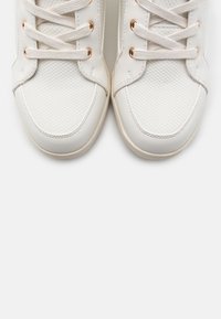 Vita sneakers med texturerade frontpaneler i tyg, vita snören som är trätt genom roséguld ögla, fotograferade ovanifrån på en enfärgad bakgrund.