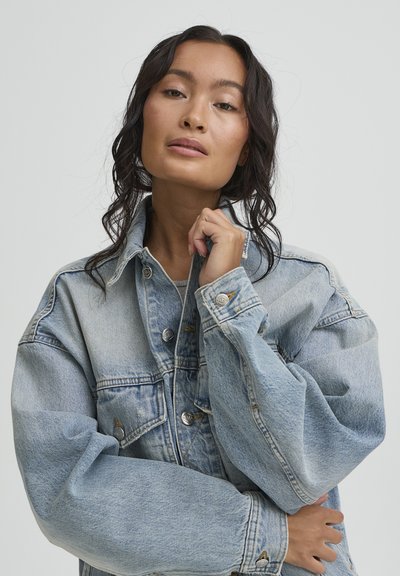 Young woman with wavy dark hair wearing oversized light blue denim jacket, holding collar with one hand and crossing arms.