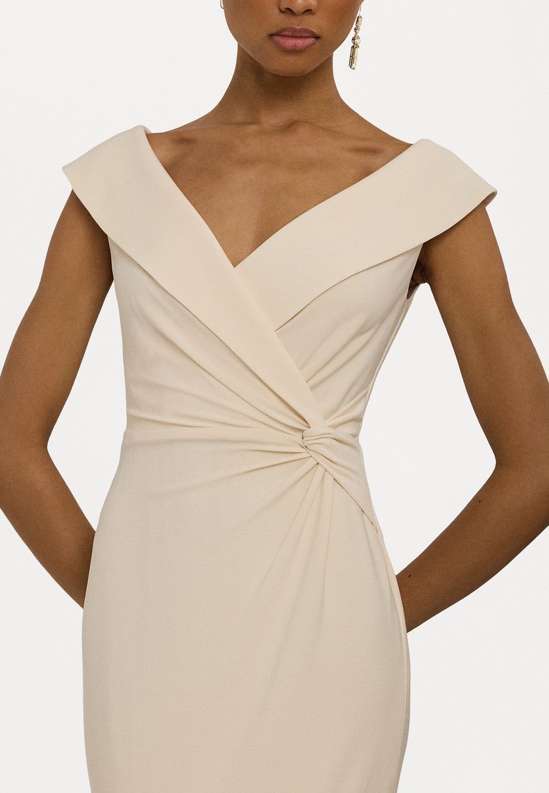 Vestido de noche beige con escote en V profundo y hombros anchos y estructurados. Presenta un detalle de cintura retorcida y textura suave y lisa.