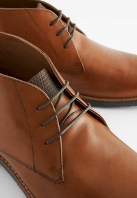 Botas de vestir de cuero marrón con un acabado liso, presentando un panel texturizado marrón y cordones grises, destacando detalles de costura simples.