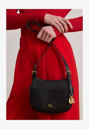 Personne en tenue rouge tenant un sac à bandoulière en cuir noir avec logo RLL doré et charms au niveau de la taille.