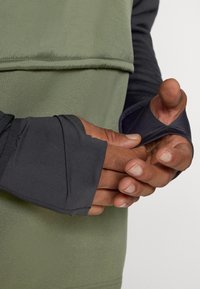 T-shirt με μακριά μανίκια, διαθέτει σκούρο γκρι σώμα και πράσινη λεπτομέρεια. Το ύφασμα είναι λείο με ελαφριά γυαλάδα. Οπές για τον αντίχειρα είναι ορατές στα μανίκια.