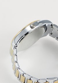 Montre en métal argenté et doré avec un bracelet poli, un boîtier rond, une lunette texturée et une couronne proéminente sur le côté.