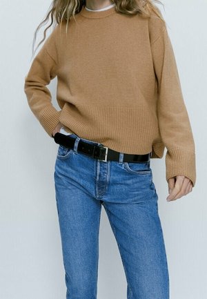 Persoon in een camelgekleurde sweater, blauwe jeans en een zwarte riem, staand tegen een effen lichte achtergrond.