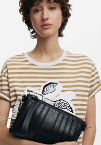 Crna kožna clutch torbica s horizontalnom rebrastom teksturom, s pletenom bijelom drškom i srebrnim hardverom, držana protiv prugaaste košulje.