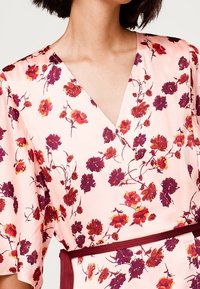 Robe enveloppante florale en tissu rose doux, ornée de motifs de fleurs violettes et rouges, avec un décolleté en V et des manches courtes, complétée par une ceinture bordeaux contrastante.