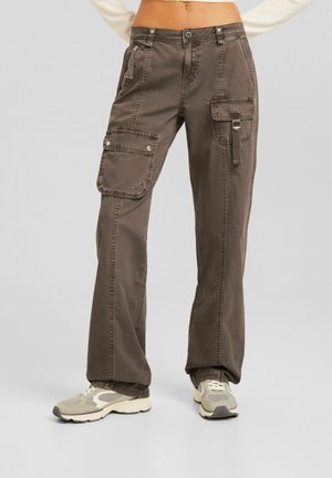 Persona que lleva pantalones cargo marrones con múltiples bolsillos y zapatillas beige, de pie frente a un fondo liso de color gris claro.