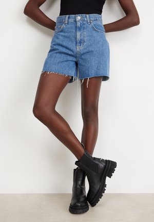 Shorts de mezclilla azul de cintura alta con dobladillo deshilachado, combinados con botas de cuero negras hasta el tobillo con suelas gruesas y paneles laterales elásticos.