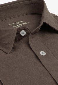 Camisa marrón de hombre de corte holgado con botones blancos, que presenta un cuello abierto y una etiqueta de marca visible en el interior del cuello.
