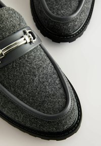 Grijze leren loafers met zwarte leren accenten, voorzien van een zilveren hardware detail over de bovenkant en een geztructureerde profielzool.