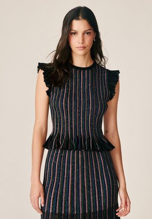 Femme portant un haut noir sans manches et une jupe à rayures verticales multicolores avec des bords froufroutants, debout devant un fond uni.