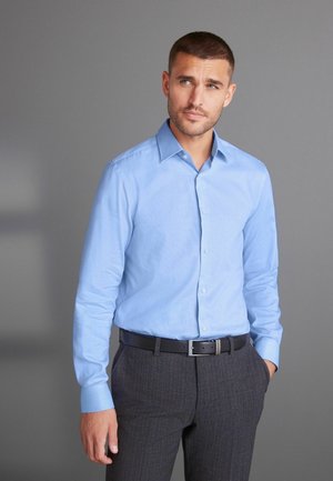 Hellblaue langärmlige Hemdbluse mit Kent-Kragen, kombiniert mit grauen gestreiften Hosen und einem schwarzen Gürtel. Glatte Textur.